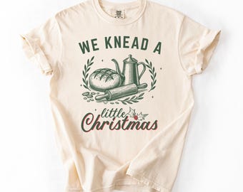May include: Cream-colored t-shirt with a green and red graphic. The graphic features a loaf of bread, a teapot, and a rolling pin, surrounded by a wreath. The text reads "WE KNEAD A little Christmas".