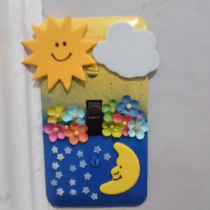 May include: A light switch cover decorated with a yellow sun, a white cloud, colorful flowers, and a yellow crescent moon on a blue background with white stars.