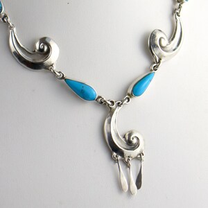 Puede incluir: Un collar de plata con piedras turquesas en forma de l&aacute;grima y adornos decorativos. El collar tiene un colgante con tres elementos colgantes de plata. La joya se exhibe sobre un maniqu&iacute; blanco.