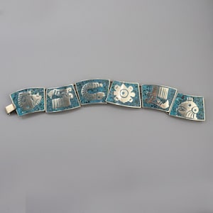Op de afbeelding: Een zilveren armband met turquoise inleg met een reeks geometrische ontwerpen. De armband bestaat uit afzonderlijke vierkante schakels, elk met een ander ontwerp.