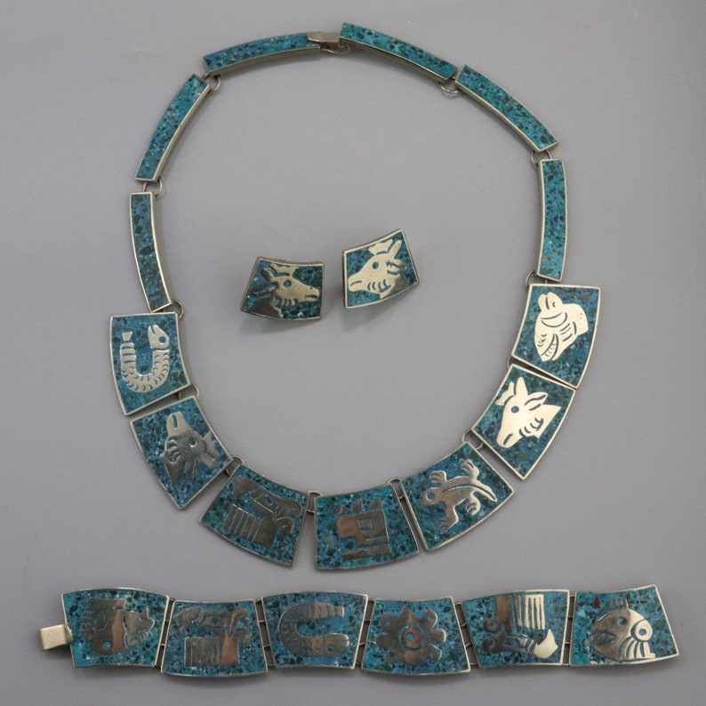 Op de afbeelding: Een turquoise en zilveren ketting- en armbandset met geometrische ontwerpen. De ketting heeft een hanger in de vorm van een hoefijzer en de armband heeft een hanger in de vorm of een vis.