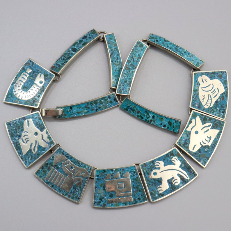 Op de afbeelding: Een turquoise en zilveren ketting met een geometrisch ontwerp. Elke schakel heeft een ander dier of symbool, waaronder een vis, een vogel, een slang en een hert. De ketting is gemaakt van sterling zilver en turquoise stenen.