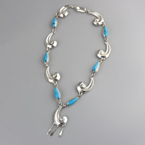 Puede incluir: Un collar plateado con una serie de formas estilizadas en espiral y piedras turquesas en forma de l&aacute;grima. El collar tiene un colgante central con tres elementos plateados colgantes. El cierre es un simple cierre de gancho y ojo.