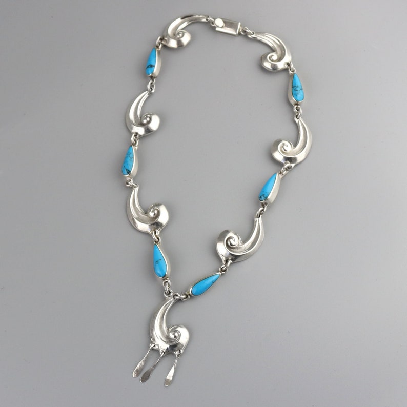 Puede incluir: Un collar plateado con una serie de formas estilizadas en espiral y piedras turquesas en forma de l&aacute;grima. El collar tiene un colgante central con tres elementos plateados colgantes. El cierre es un simple cierre de gancho y ojo.