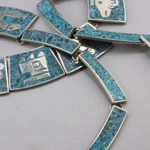 Op de afbeelding: Een turquoise en zilveren ketting met geometrische ontwerpen. De ketting is gemaakt van meerdere gekoppelde stukken, elk met een ander ontwerp. De turquoise is een levendige blauwgroene kleur en het zilver is gepolijst.