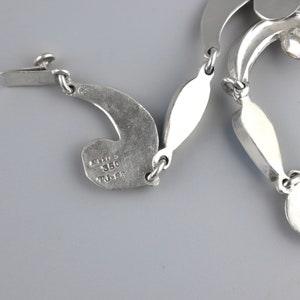 Puede incluir: Un collar plateado con un dise&ntilde;o &uacute;nico y fluido. El collar presenta eslabones curvos en forma de hoja conectados por peque&ntilde;os anillos. El cierre es visible a la izquierda, y el collar est&aacute; estampado con "Mexico 350 TJ-62".