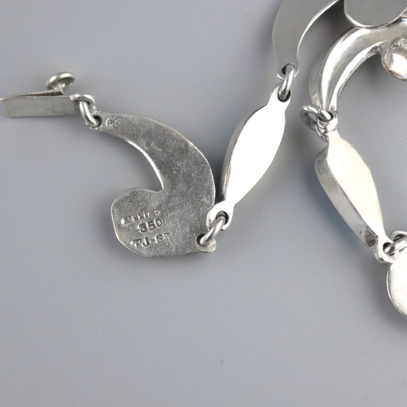 Puede incluir: Un collar plateado con un dise&ntilde;o &uacute;nico y fluido. El collar presenta eslabones curvos en forma de hoja conectados por peque&ntilde;os anillos. El cierre es visible a la izquierda, y el collar est&aacute; estampado con "Mexico 350 TJ-62".