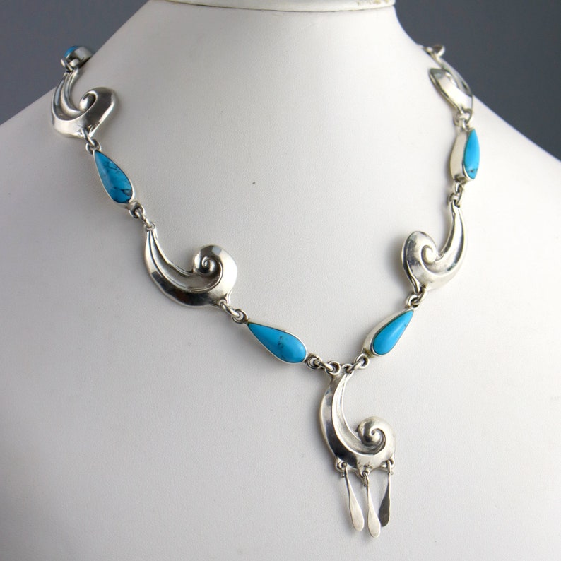 Puede incluir: Un collar de plata con piedras turquesas en forma de l&aacute;grima y adornos decorativos. El collar tiene un colgante con tres elementos colgantes de plata. La joya se exhibe sobre un maniqu&iacute; blanco.