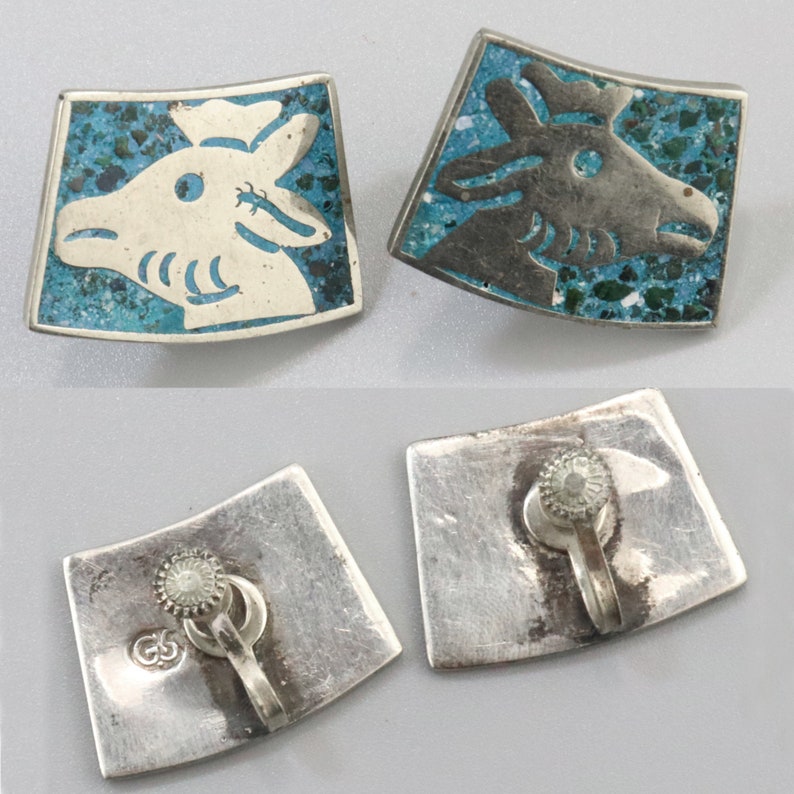 Op de afbeelding: Een paar zilveren oorbellen met turquoise en wit emaille-ontwerpen. De oorbellen hebben een gestileerd dierenkopontwerp. De achterkant van de oorbellen is zilver met een clip sluiting.