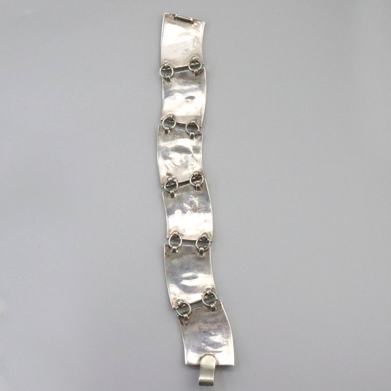 Op de afbeelding: Een zilveren armband met een golvend ontwerp en meerdere verbonden schakels. Elke schakel heeft een getextureerd oppervlak en een klein rond detail.