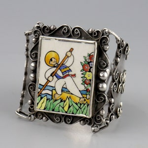 Könnte beinhalten: Ein silbernes Armreif-Armband mit einem rechteckigen Keramikfliesen-Mittelteil. Die Fliese zeigt eine farbenfrohe Illustration einer Person in einem Boot, mit Blumen auf der rechten Seite. Das Armband hat dekorative Schnörkel und Kugelakzente.