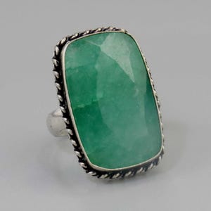Puede incluir: Anillo de plata con una gran piedra preciosa verde rectangular. La piedra preciosa está facetada y engastada en un marco de plata con un diseño de cuerda trenzada. Una joya llamativa.