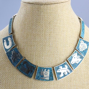 Op de afbeelding: Een turquoise en zilveren ketting met geometrische ontwerpen. Elke schakel heeft een ander ontwerp, waaronder een hoefijzer, een vis en een vogel. De ketting is gemaakt van metaal en heeft een blauw en wit kleurenschema.