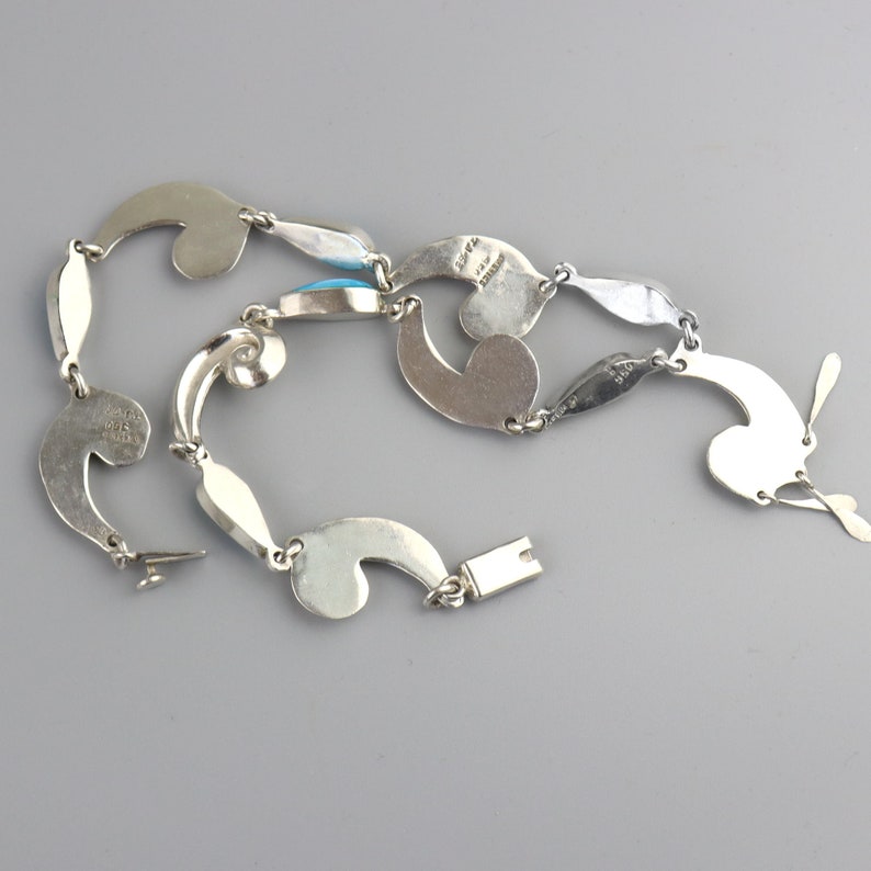 Puede incluir: Un collar de plata con un dise&ntilde;o &uacute;nico y fluido. El collar presenta formas estilizadas interconectadas, incluyendo elementos curvos y eslabones en forma de l&aacute;grima. Incorpora peque&ntilde;as piedras turquesas ovaladas. El cierre es de tipo gancho y barra.