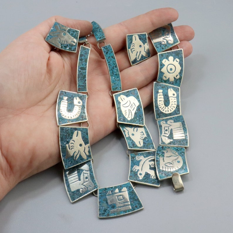 Op de afbeelding: Een turquoise en zilveren ketting met een reeks rechthoekige hangers met verschillende dierlijke en geometrische ontwerpen.
