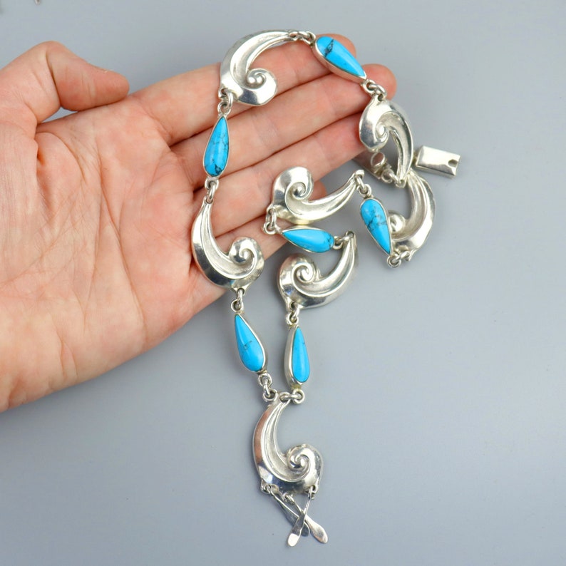 Puede incluir: Un collar de plata con piedras turquesas en forma de l&aacute;grima y dise&ntilde;os de olas estilizadas y onduladas. El collar est&aacute; compuesto por m&uacute;ltiples secciones enlazadas, con las piedras turquesas intercaladas en el dise&ntilde;o. El cierre es visible en la parte superior.