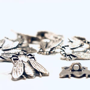 May include: Close-up of several silver-colored fly-shaped metal embellishments. The detailed insect designs feature wings, bodies, and legs. One embellishment has a loop on the back. The background is white.