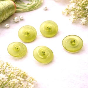 May include: Five pale yellow, spiral-shaped buttons with a translucent appearance. The buttons are arranged on a white surface, with green thread and small white pearls in the background. Delicate white flowers are also visible.