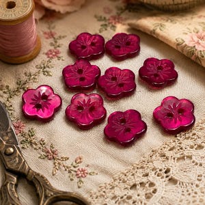 May include: A collection of nine translucent, deep magenta, flower-shaped buttons. Each button features a five-petal design with a central indentation and two holes for sewing. The buttons are arranged in a cluster, showcasing their glossy finish.