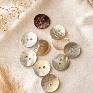 May include: A collection of ten round buttons with two holes each. Nine buttons feature a smooth, iridescent, off-white and light brown pearlescent finish, while one button is a dark, textured brown with a marbled appearance and a glossy surface.