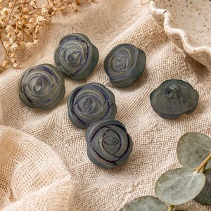 May include: Six gray rose-shaped decorative objects with blue accents are arranged on a beige fabric. The objects have a detailed floral design. Dried flowers and greenery are in the background.