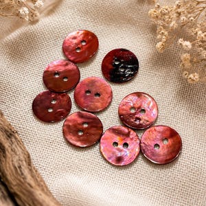May include: A collection of eight circular buttons featuring a deep reddish-brown hue with a pearlescent, iridescent finish. Each button is crafted with two central holes for attachment, displaying a smooth, reflective surface with natural variations.