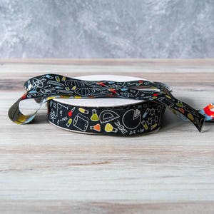 May include: Black and white ribbon with a science theme. The ribbon is printed with images of beakers, test tubes, atoms, and other science-related symbols. The ribbon is rolled up on a white spool.