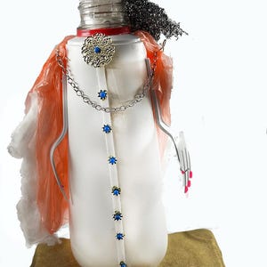 May include: A quirky sculpture made from recycled items. It features a white bottle body with blue embellishments, a blue hat, and a red collar. The figure has a chain necklace and a fork arm with pink painted nails. It stands on a brown base.