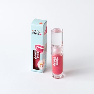 Peut inclure: Un tube de brillant à lèvres transparent avec une teinte rose et un bouchon transparent, à côté d'une boîte bleu clair. La boîte présente le texte "Pama Dama" et un dessin de lèvres. Le brillant à lèvres est étiqueté "So Juicy Gloss".