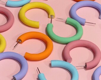 May include: A collection of colorful hoop earrings in various pastel shades, including pink, blue, green, yellow, and orange. The earrings are made of a thick, rounded material and have a simple, modern design.