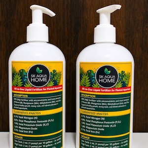May include: Two white plastic pump bottles of SK Aqua Home all-in-one liquid fertilizer for planted aquariums. The labels feature a green leaf pattern, the brand name, a description of NPK fertilizer with micronutrients, and dosage instructions.