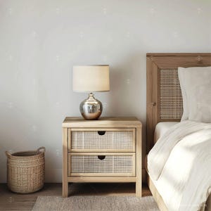 May include: A wooden nightstand with two drawers featuring a woven rattan front. The nightstand has a lamp with a white shade and a silver base. The nightstand is next to a bed with a white headboard and white bedding.