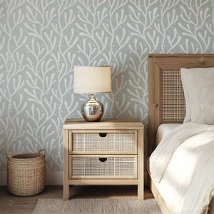 May include: A light blue wallpaper with a white branch pattern. A wooden nightstand with two drawers has a lamp on top. A white bed with a wooden headboard is in the background.