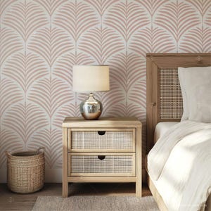 May include: A wooden nightstand with two drawers and a woven rattan front. A lamp with a white shade sits on top of the nightstand. The nightstand is in front of a wall with a pink and white palm leaf patterned wallpaper.