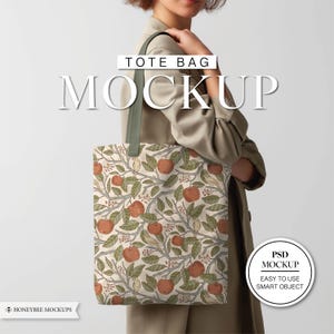 Könnte beinhalten: Tote Bag Mockup mit cremefarbenem Hintergrund und einem sich wiederholenden Muster aus Äpfeln, Blättern und Vögeln. Die Tasche hat grüne Träger und die Worte "TOTE BAG MOCKUP" sind oben aufgedruckt. Die Tasche wird von einer Person in einem beigen Mantel gehalten.