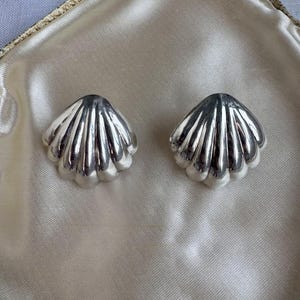May include: A pair of silver seashell-shaped earrings. Each earring has a scalloped edge and vertical ridges, reflecting light. The earrings are displayed on a satin-like, off-white surface.