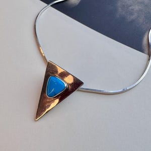 Op de afbeelding: Een zilverkleurige slangenketting met een grote, gepolijste driehoekige hanger. De hanger heeft een glad, reflecterend oppervlak en is in het midden bezet met een levendige blauwe, afgeronde driehoekige steen.
