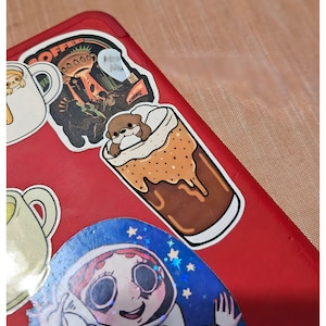 May include: A collection of colorful stickers on a red surface. The stickers include a cartoon otter in a coffee drink, a space-themed illustration, and a mug with a cartoon character. The stickers are vibrant and detailed.