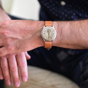 Può includere: Un orologio da polso vintage con cassa color argento e cinturino in pelle marrone chiaro. Il quadrante dell'orologio è color crema con numeri e lancette nere. L'orologio è indossato al polso.