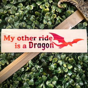 Può includere: Un adesivo rettangolare con sfondo color crema che riporta il testo "My other ride is a Dragon" in un carattere sfumato rosso, arancione e viola, accompagnato da una grafica stilizzata della sagoma di un drago rosso.