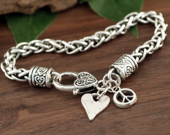 May include: A silver chain bracelet with a heart charm, a peace sign charm, and a heart-shaped clasp. The bracelet is on a wooden surface.