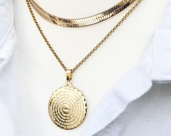 May include: A gold chain necklace with a round pendant featuring a circular inscription. The pendant is attached to a thinner gold chain. The necklace is layered over a thicker gold chain.