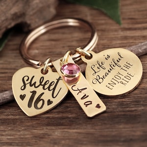 May include: Gold-tone keychain with a heart-shaped charm that says "Sweet 16" and a round charm that says "Life is Beautiful Enjoy the Ride". There is also a small rectangular charm with the name "Ava" and a pink rhinestone.