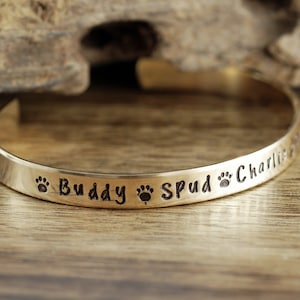 May include: Gold metal bracelet with engraved paw prints and the names "Buddy", "Spud", and "Charlie".