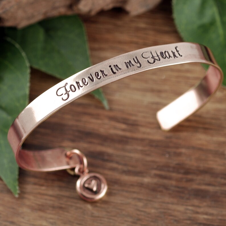 Puede incluir: Una pulsera de cobre con un dije en forma de coraz&oacute;n. La pulsera est&aacute; grabada con las palabras "Forever in my Heart".