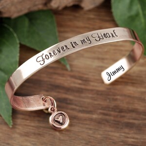 Puede incluir: Pulsera de brazalete de oro rosa con la inscripci&oacute;n "Forever in my Heart" y un nombre personalizado "Jimmy". La pulsera tiene un peque&ntilde;o dije de coraz&oacute;n.