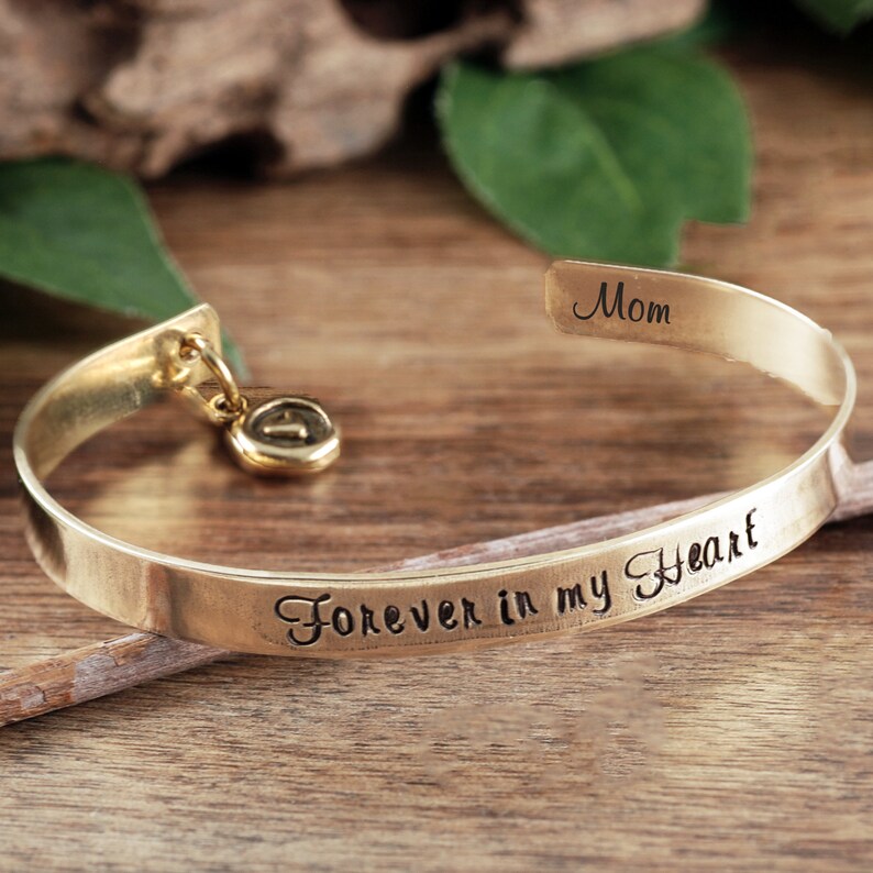 Puede incluir: Pulsera de brazalete de oro con las palabras "Forever in my Heart" grabadas. La pulsera tiene un peque&ntilde;o dije con el n&uacute;mero "1". La parte posterior de la pulsera est&aacute; grabada con la palabra "Mam&aacute;".