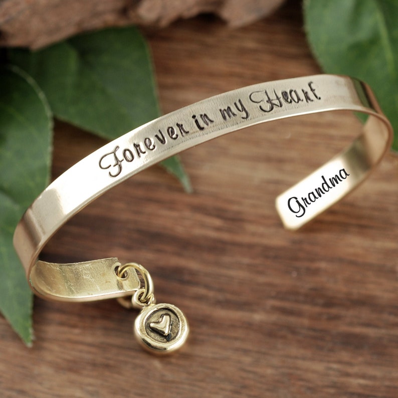 Puede incluir: Pulsera dorada con un dije en forma de coraz&oacute;n. La pulsera est&aacute; grabada con las palabras "Forever in my Heart" y "Grandma".