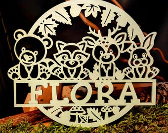 Op de afbeelding: Een houten uitsnijding van een cirkel met een beer, een wasbeer, een hert en een konijn. De dieren zitten allemaal en kijken naar voren. Het woord "FLORA" is onder de dieren uitgesneden. De uitsnijding bevindt zich op een bruin houten oppervlak.