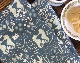 Puede incluir: Un paño de cocina de tela azul oscuro con un patrón repetitivo de caballitos de mar de color verde claro, coral blanco, algas y pequeñas burbujas circulares. El tejido tiene una textura tejida y muestra un denso motivo marino.
